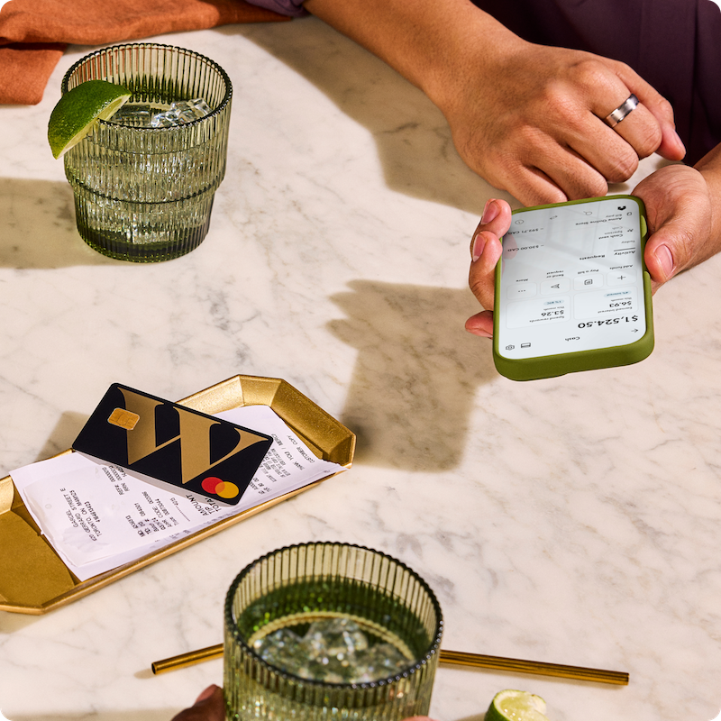 Person sitting at marble tabletop in restaurant with Wealthsimple app open on their phone and prepaid Mastercard out for payment.