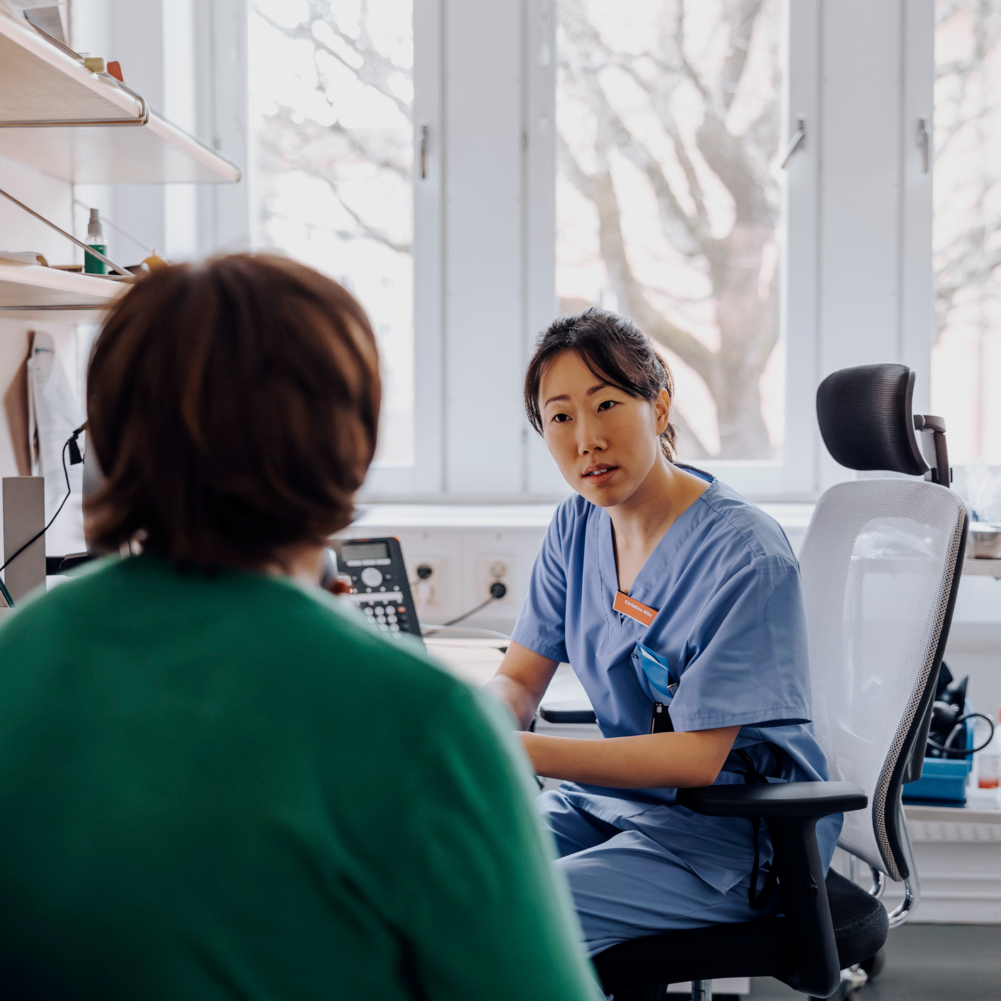 A female doctor sits in blue scrubs in front of a patient
