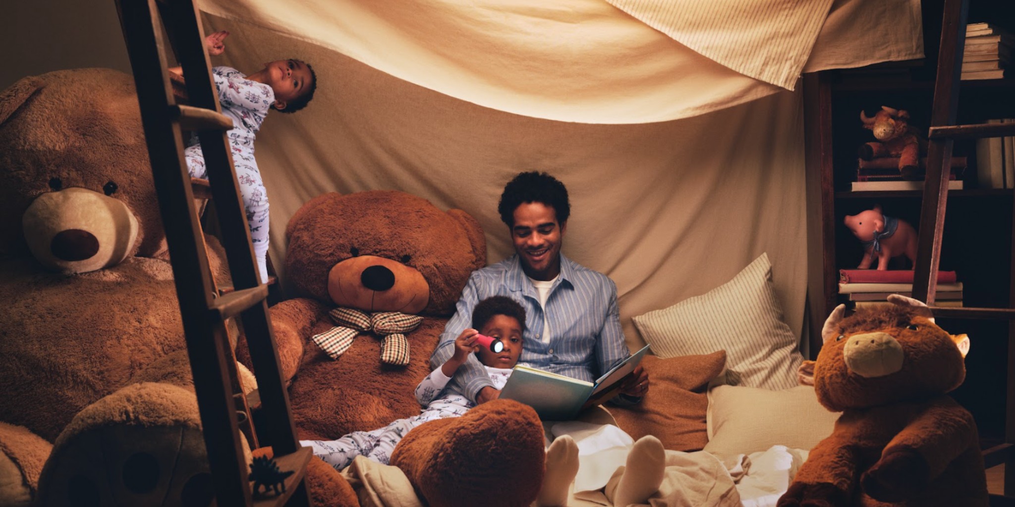 A man sits in a pillow fort reading a book with his kids