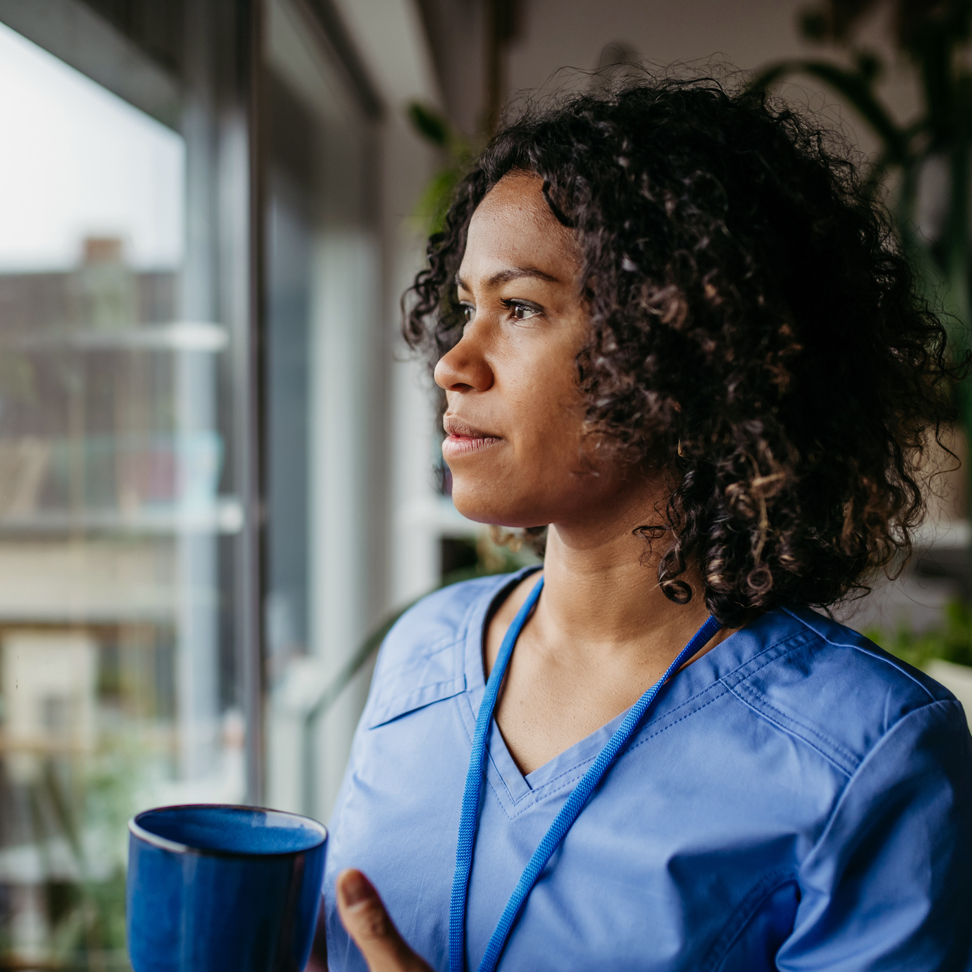 Woman in blue scrubs looking out window holding a coffee cup