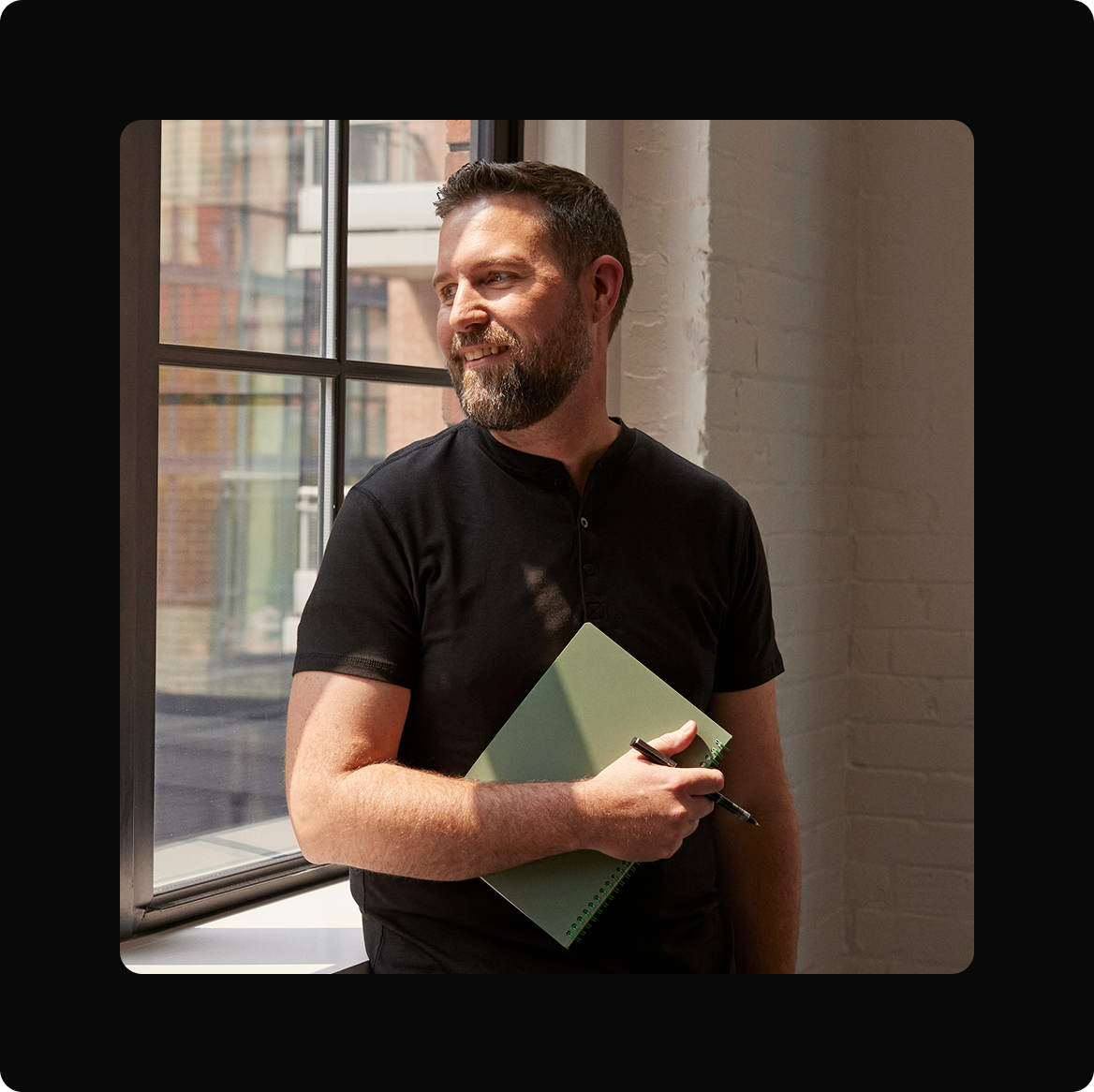 Professional man standing by a sunlit window, smiling while holding a green notebook and pen.